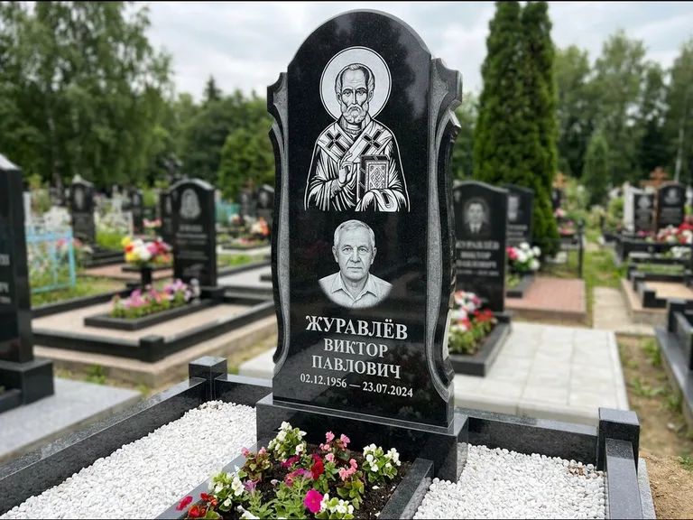 Памятник с гравировкой иконы: стела с образом в полный рост на обороте — гравировка образа Матроны Московской, низкие кусты спиреи в цвету, работа «Лебедева» в Мурманске