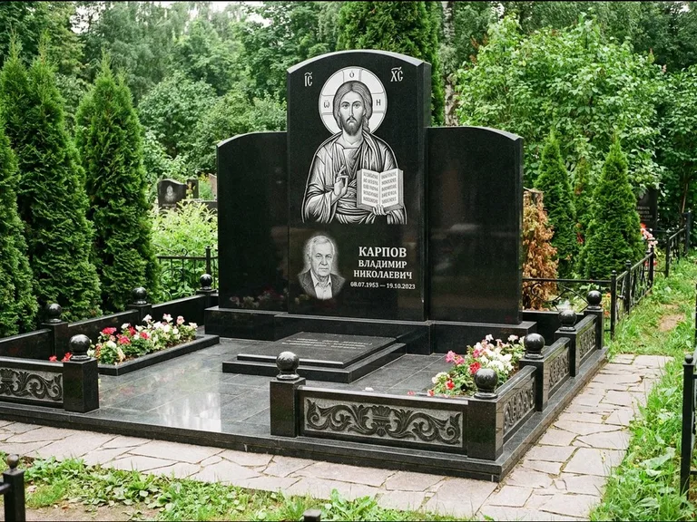 Памятник с гравировкой иконы: широкая панорамная стела с золочёным нимбом — гравировка образа Матроны Московской, цветник с белыми хризантемами, работа «Мельникова» в Мурманске
